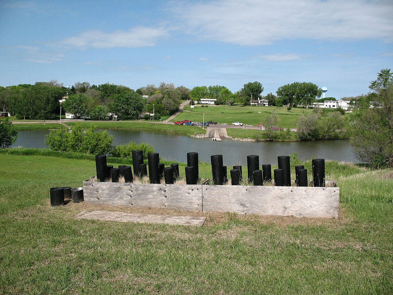 A row of black fireworks launch tubes stands behind a low wooden barrier beside a lake.