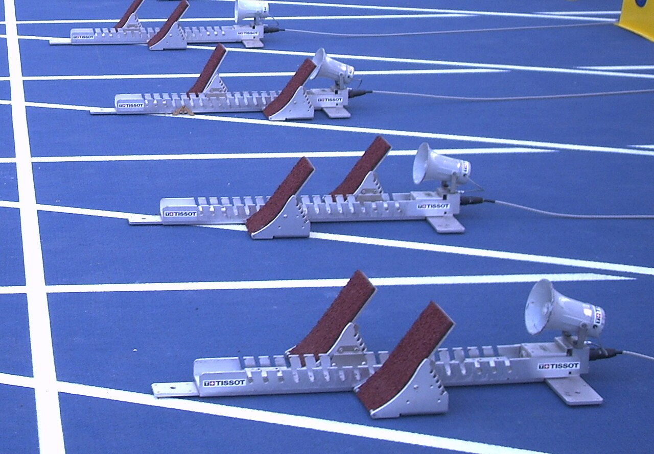Four sets of starting blocks are positioned in separate lanes at a track starting line.